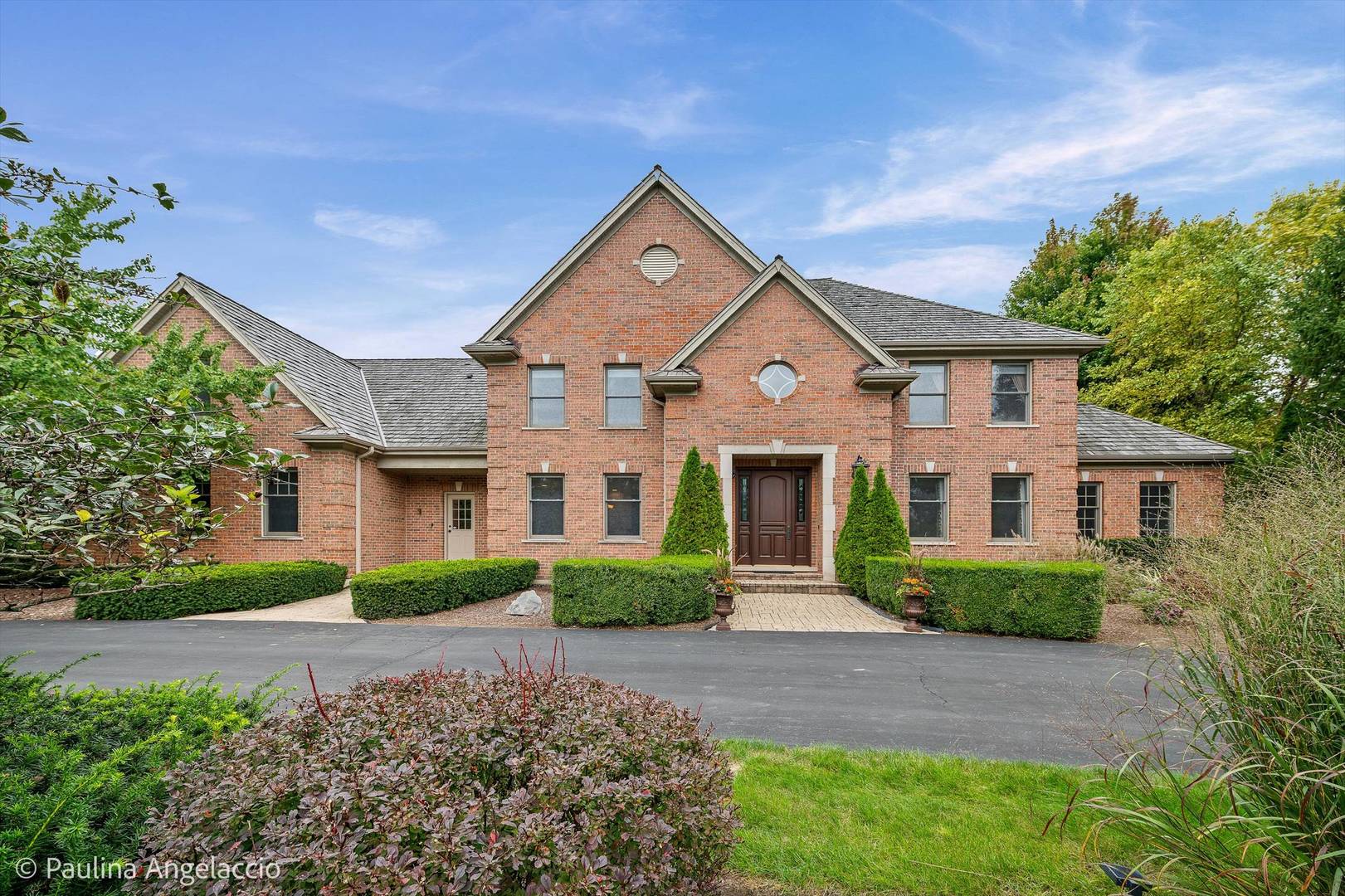 Two-story red brick home with a gabled roof, trimmed hedges, and a paved driveway.