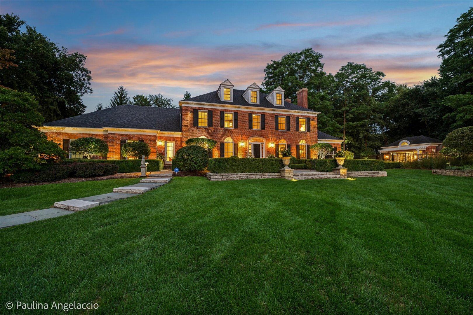 Red-brick home with illuminated windows, dormers, landscaped grounds, and a broad lawn at dusk.