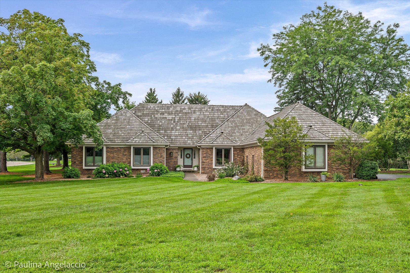 Single-story brick home with a gray shingle roof, landscaped garden, and broad green lawn.