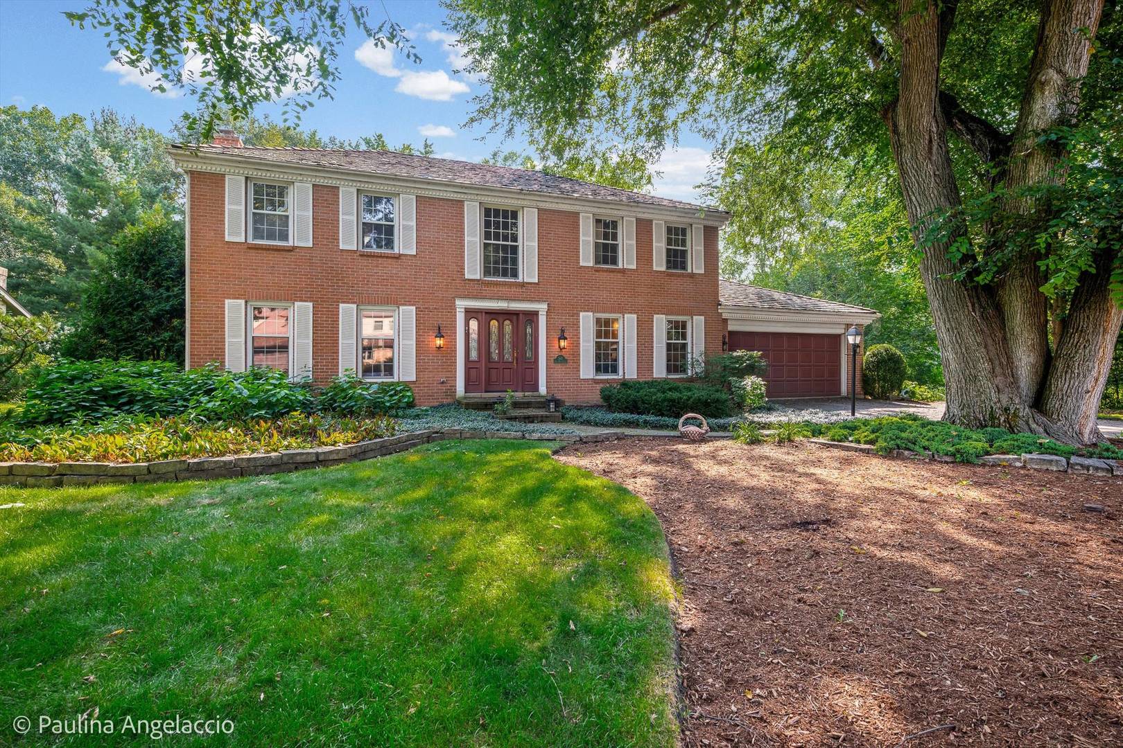Two-story red brick home with white shutters, attached garage, and landscaped lawn beneath mature trees.