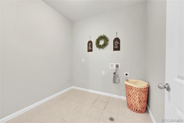 Empty utility room with wall hookups, beige tile flooring, and a woven laundry hamper.