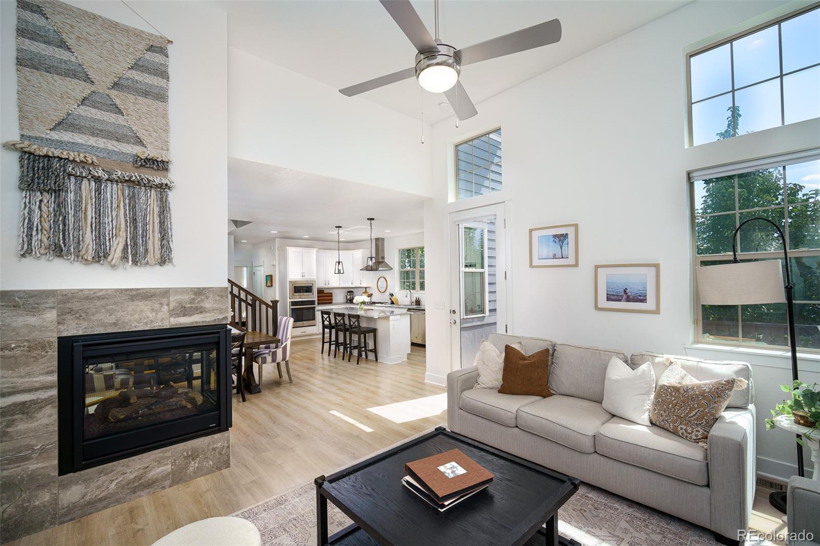 Living room with a tiled fireplace, vaulted ceiling, large windows, and an open kitchen.