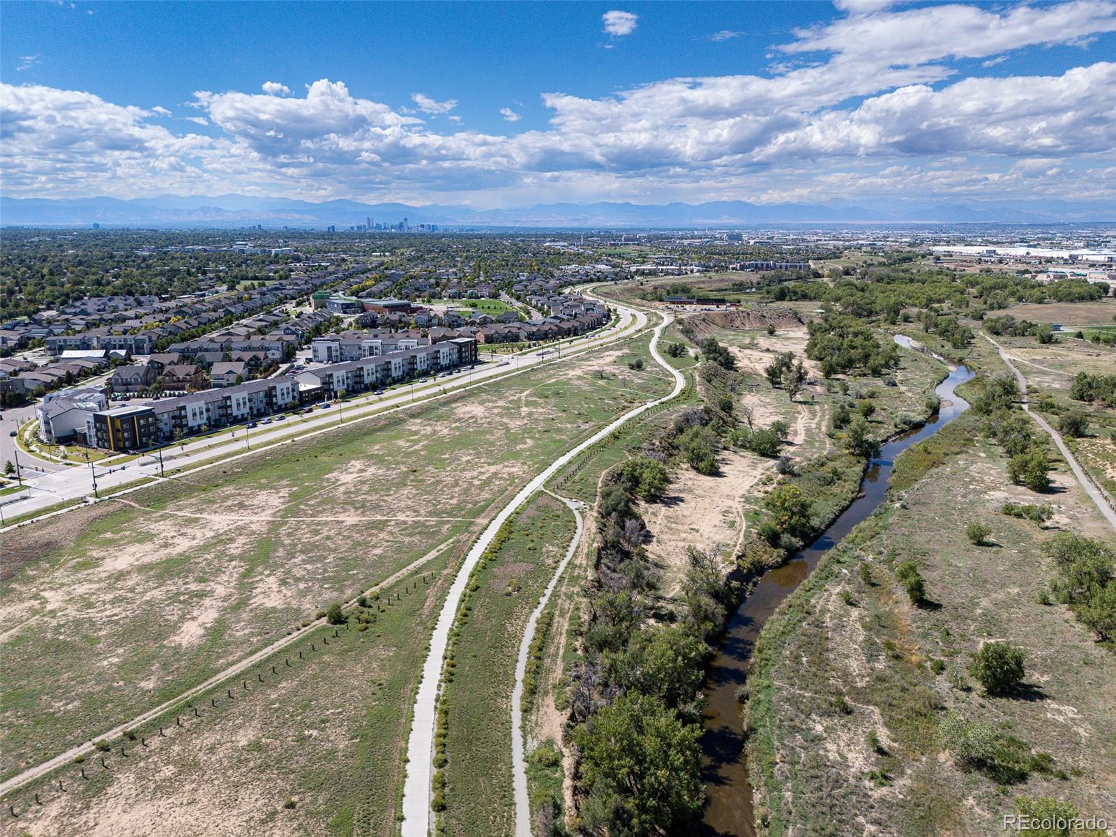 Aerial view of Denver neighborhoods and a winding creek, with the skyline and Rocky Mountains beyond.