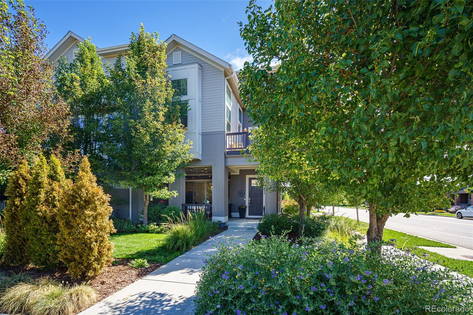 Gray two-story home with a covered entry and balcony, framed by leafy trees and landscaped gardens.
