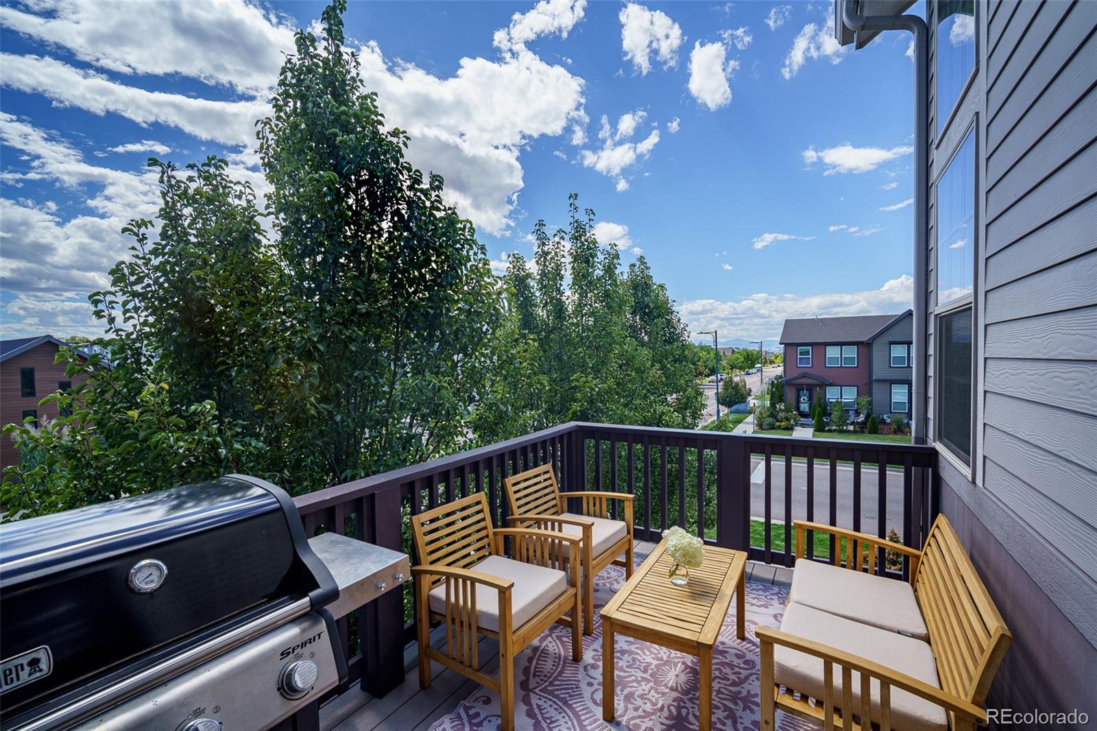 Furnished balcony with wooden seating and a grill overlooking trees and neighboring homes.