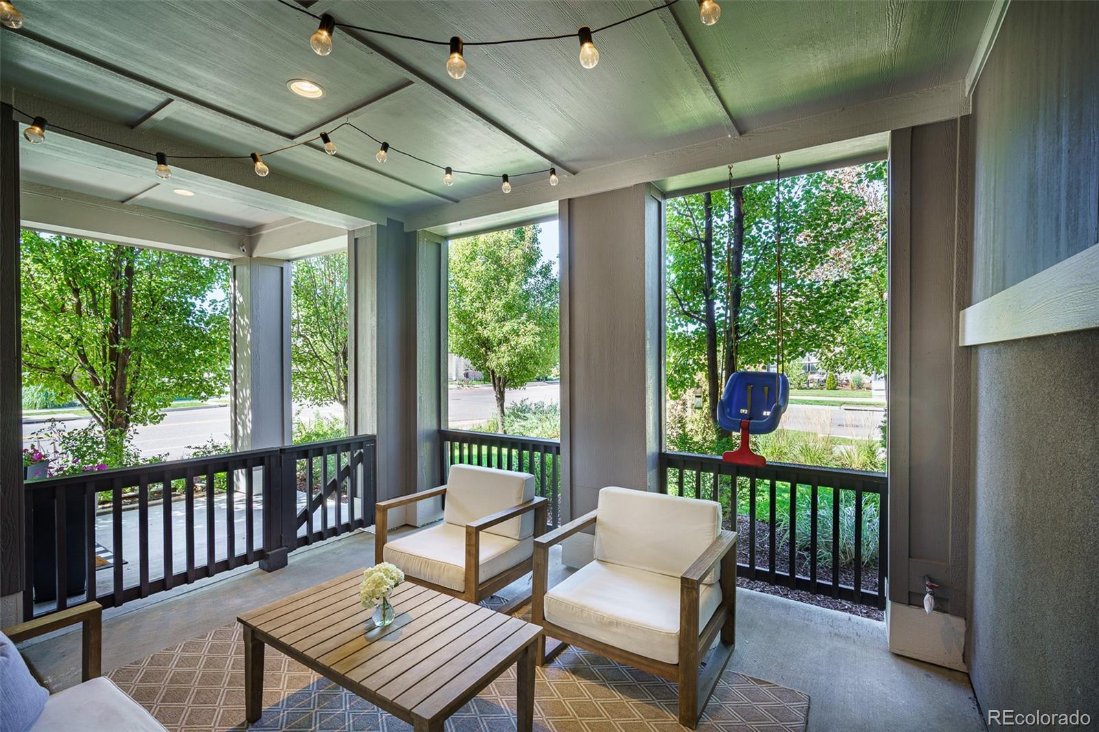 Covered porch with cushioned chairs, a wooden coffee table, string lights, and leafy views.