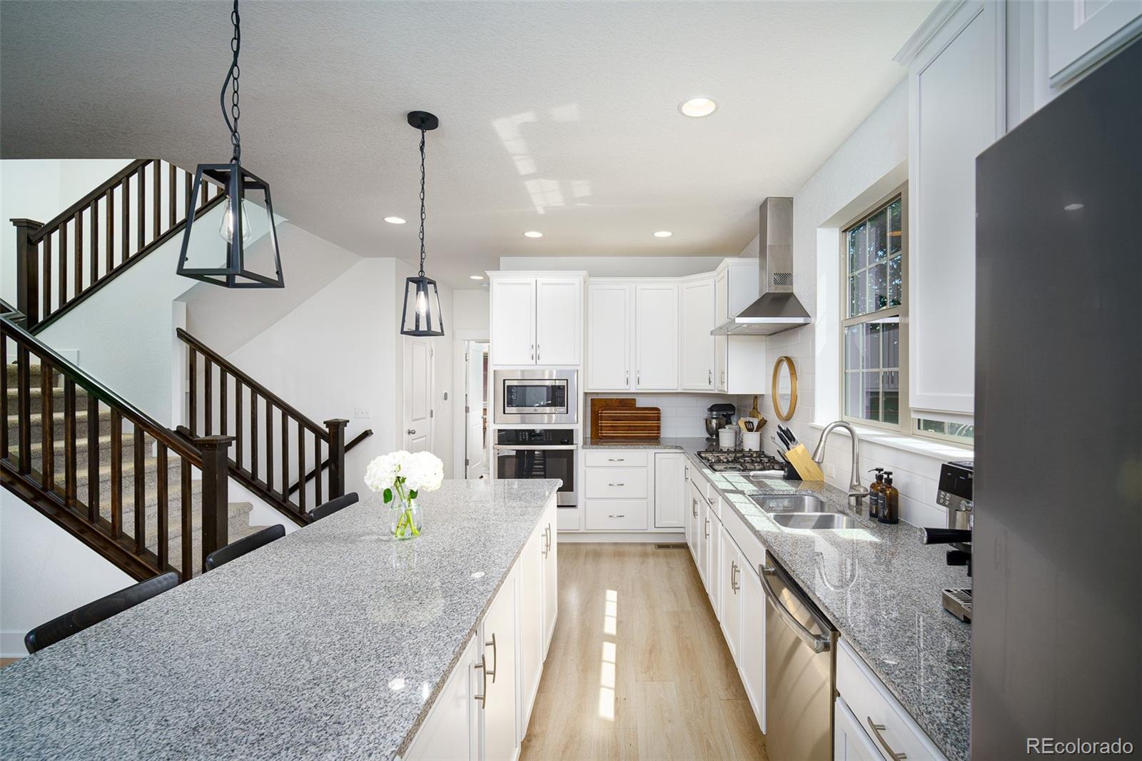 Bright kitchen with a granite island, white cabinets, stainless steel appliances, and dark wood staircase railing.