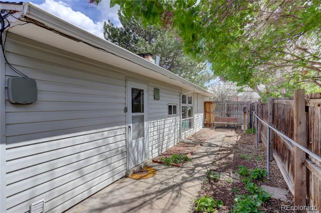 Side of a single-story home beside a concrete walkway and wooden fence under a leafy tree.