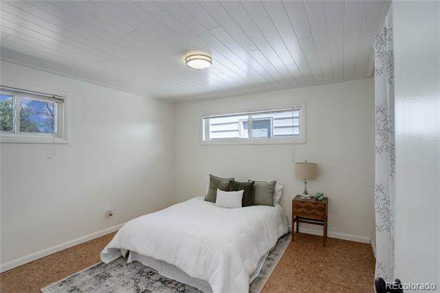Staged bedroom with a white bed, cork flooring, two small windows, and a wooden bedside table.