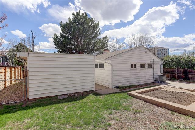 Light-colored house and shed in a fenced backyard, with a raised garden bed and large pine tree.