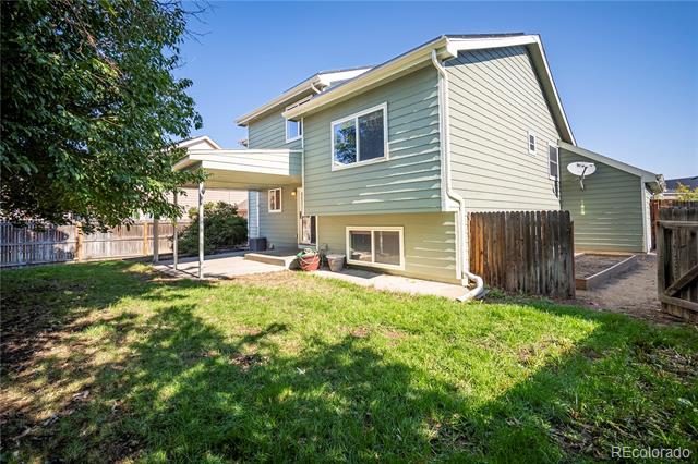 Home’s rear exterior with sage-green siding, covered patio, fenced lawn, and mature shade tree.