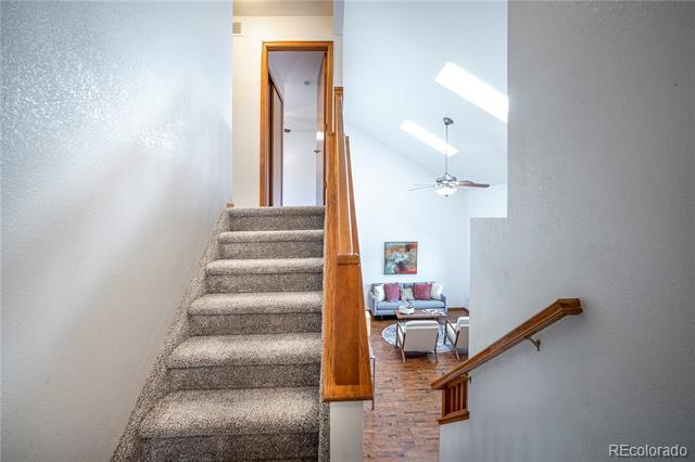 Carpeted staircase with a wooden railing overlooks a furnished living room beneath skylights.