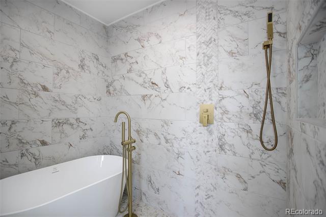 Bathroom with a freestanding tub, gold-toned fixtures, and marble-look tiled walls.