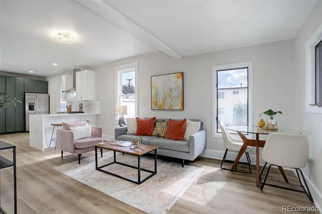 Staged living room with gray sofa, pink armchair, and dining nook beside a dark green kitchen.