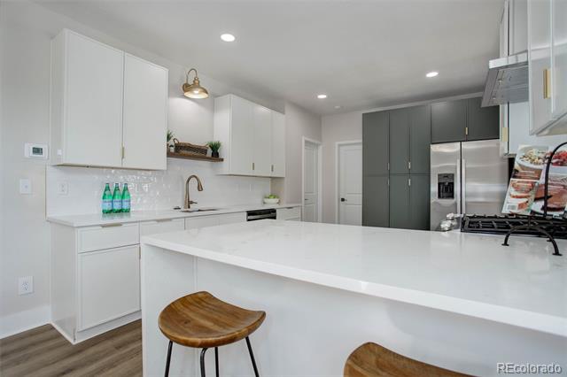 Kitchen with a large white island, wooden stools, dark gray cabinets, and stainless steel refrigerator.