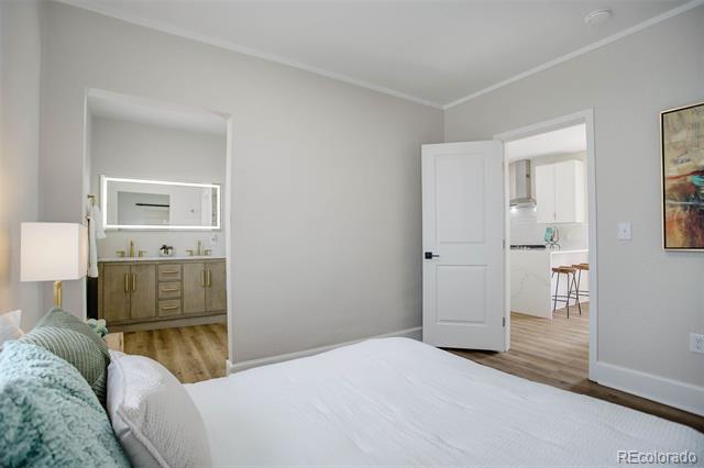 Bedroom with a white bed and teal pillows, adjoining a double-sink vanity and bright room with bar stools.