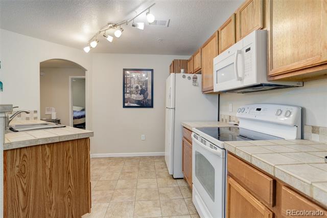 Kitchen with wood cabinets, tiled counters, and white appliances; an arched doorway leads to another room.