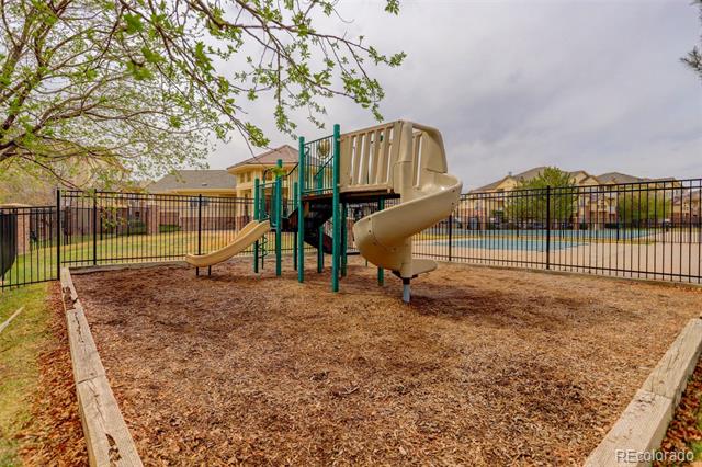 Fenced playground with a tan spiral slide, wood-chip ground, and pool beyond.