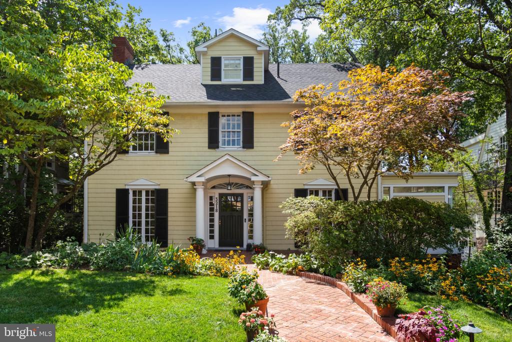 Pale yellow two-story house with black shutters, a brick garden path, and mature trees.