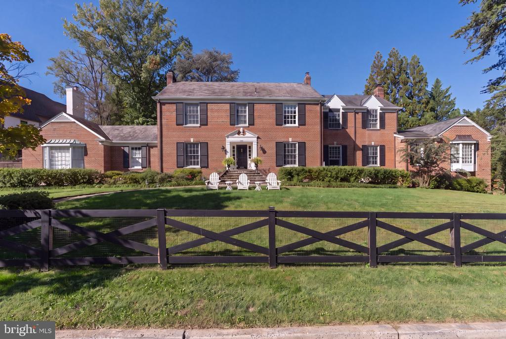 Red-brick Colonial home with black shutters, white patio chairs, and a broad lawn behind a dark wooden fence.