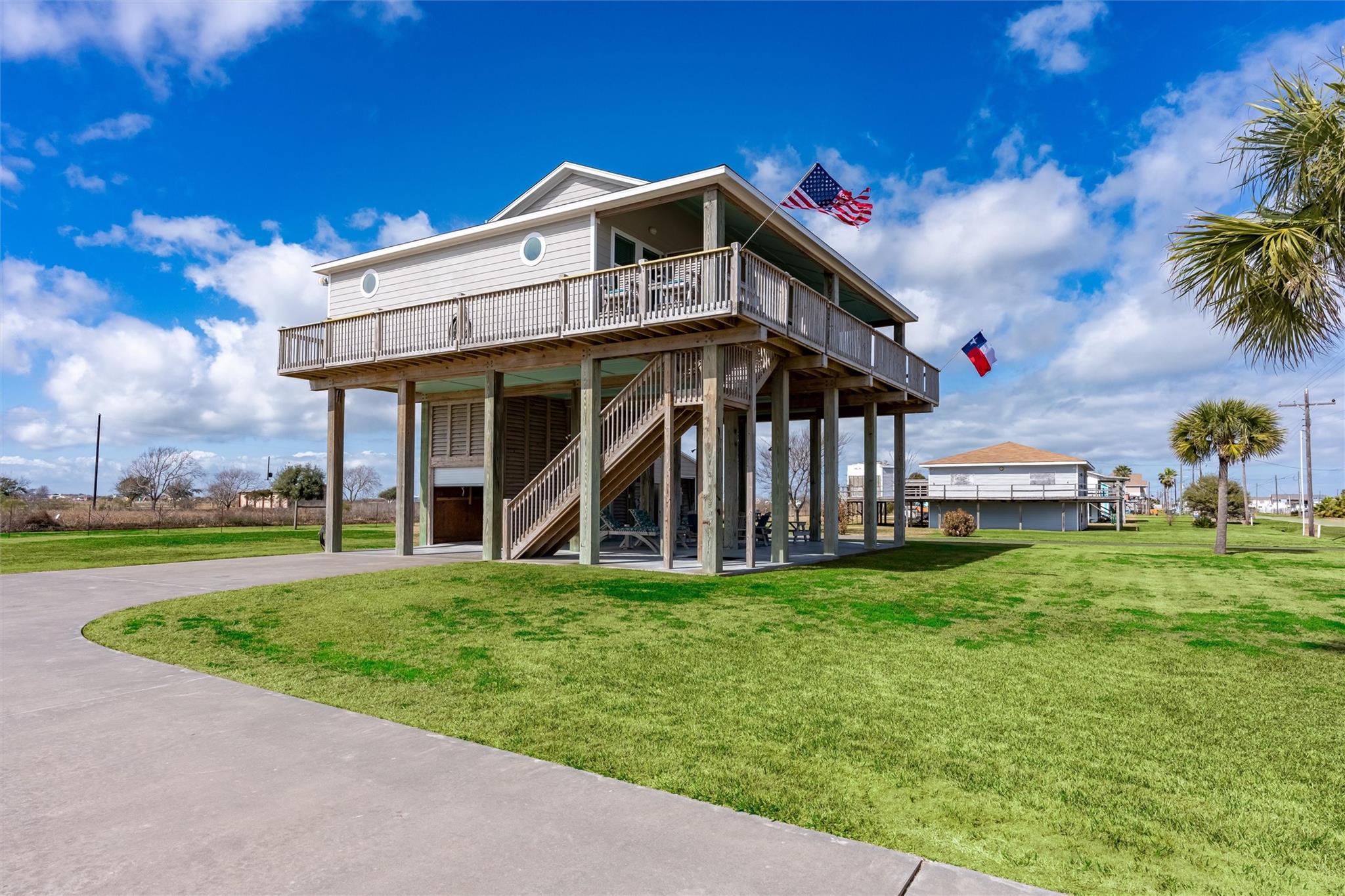 Elevated coastal house with a wraparound deck, exterior stairs, round windows, and American and Texas flags.