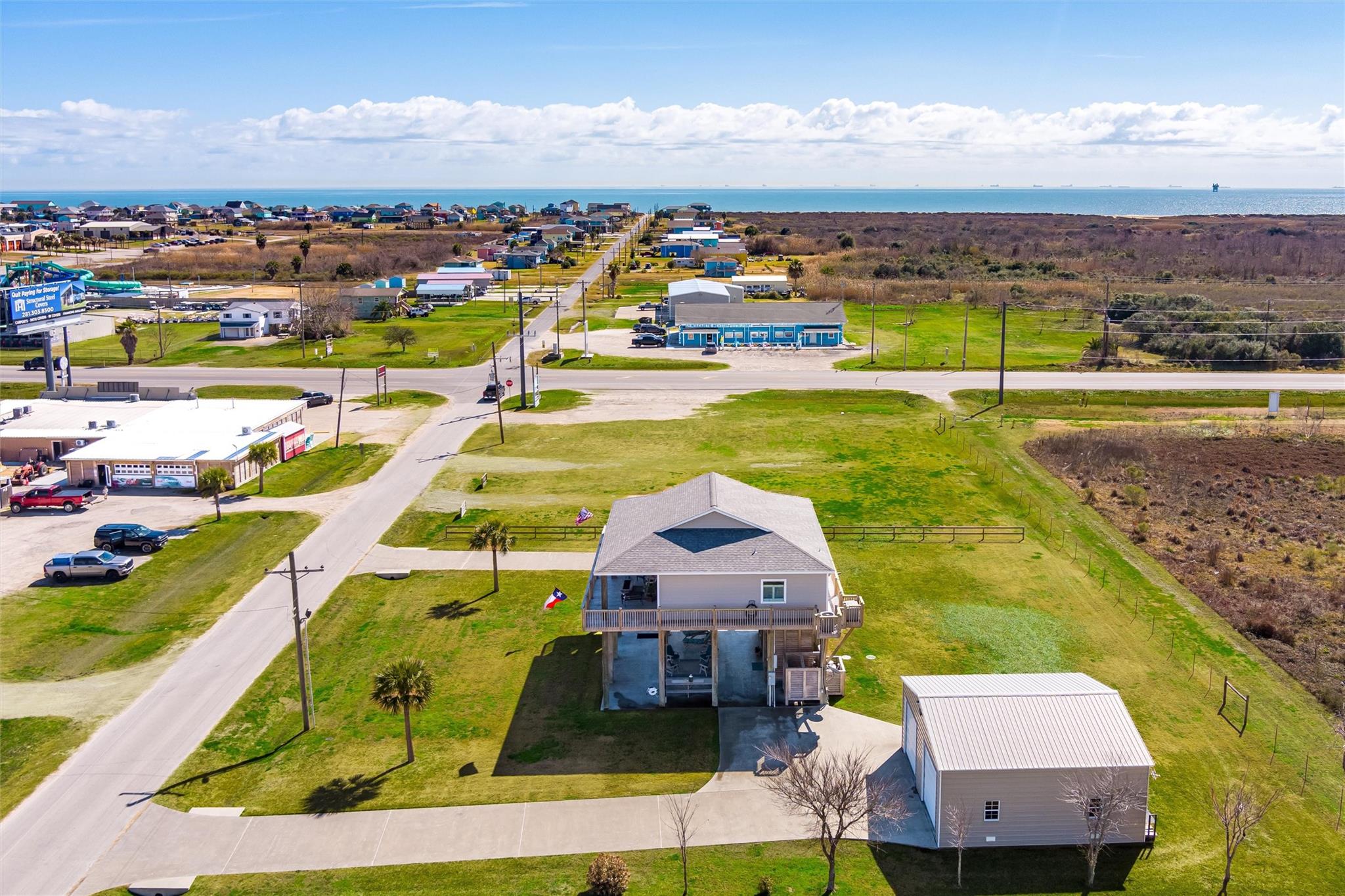 Raised coastal house and detached outbuilding on a grassy lot, with the Gulf shoreline in the distance.