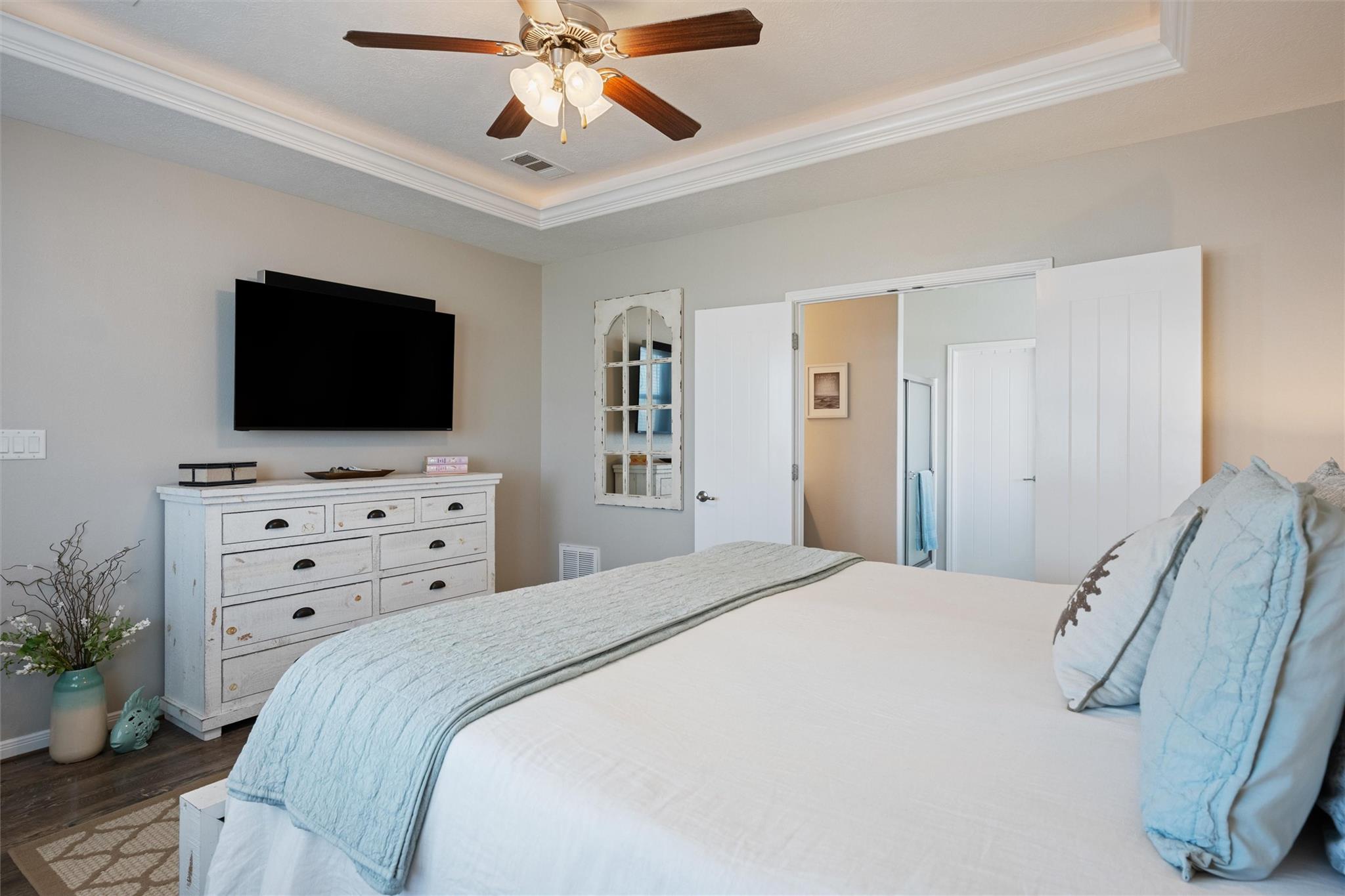 Furnished bedroom with a white bed, pale blue bedding, dresser, wall-mounted TV, and ceiling fan.
