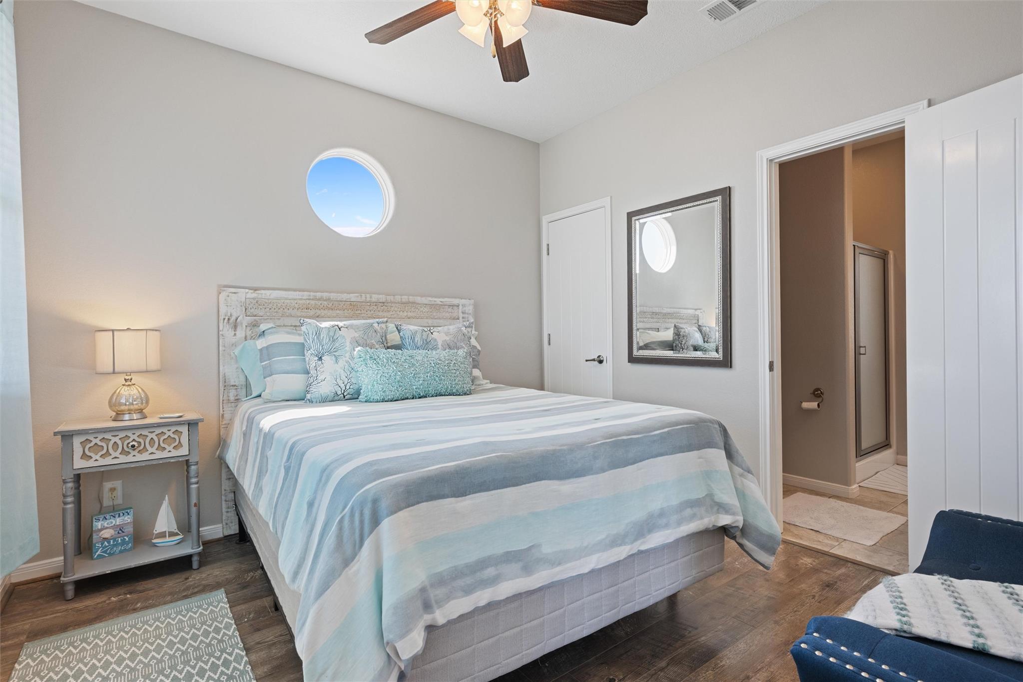 Furnished bedroom with blue striped bedding, a round window, and an open bathroom doorway.