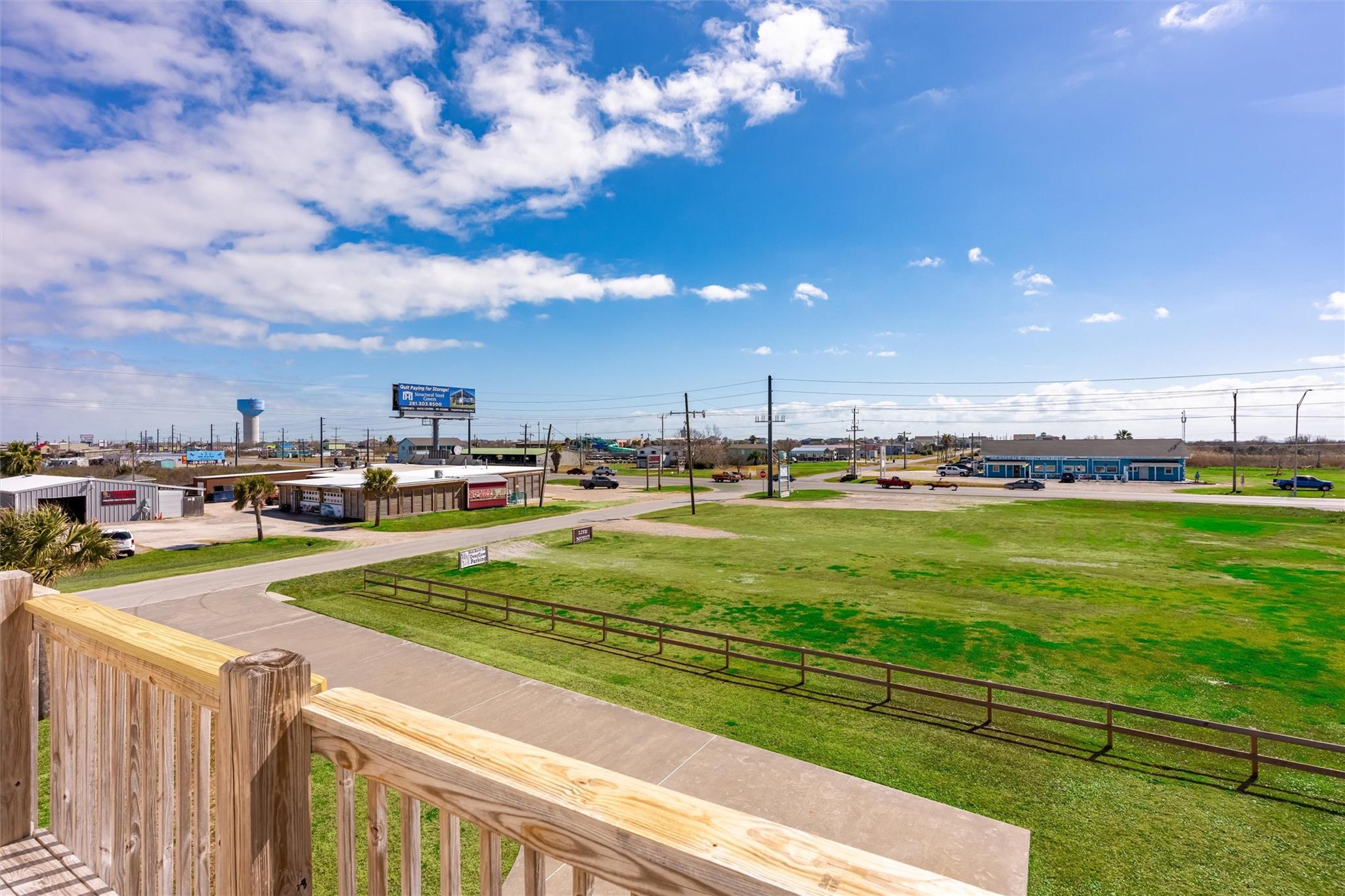 View from a wooden deck across a grassy lot toward nearby businesses and a water tower.
