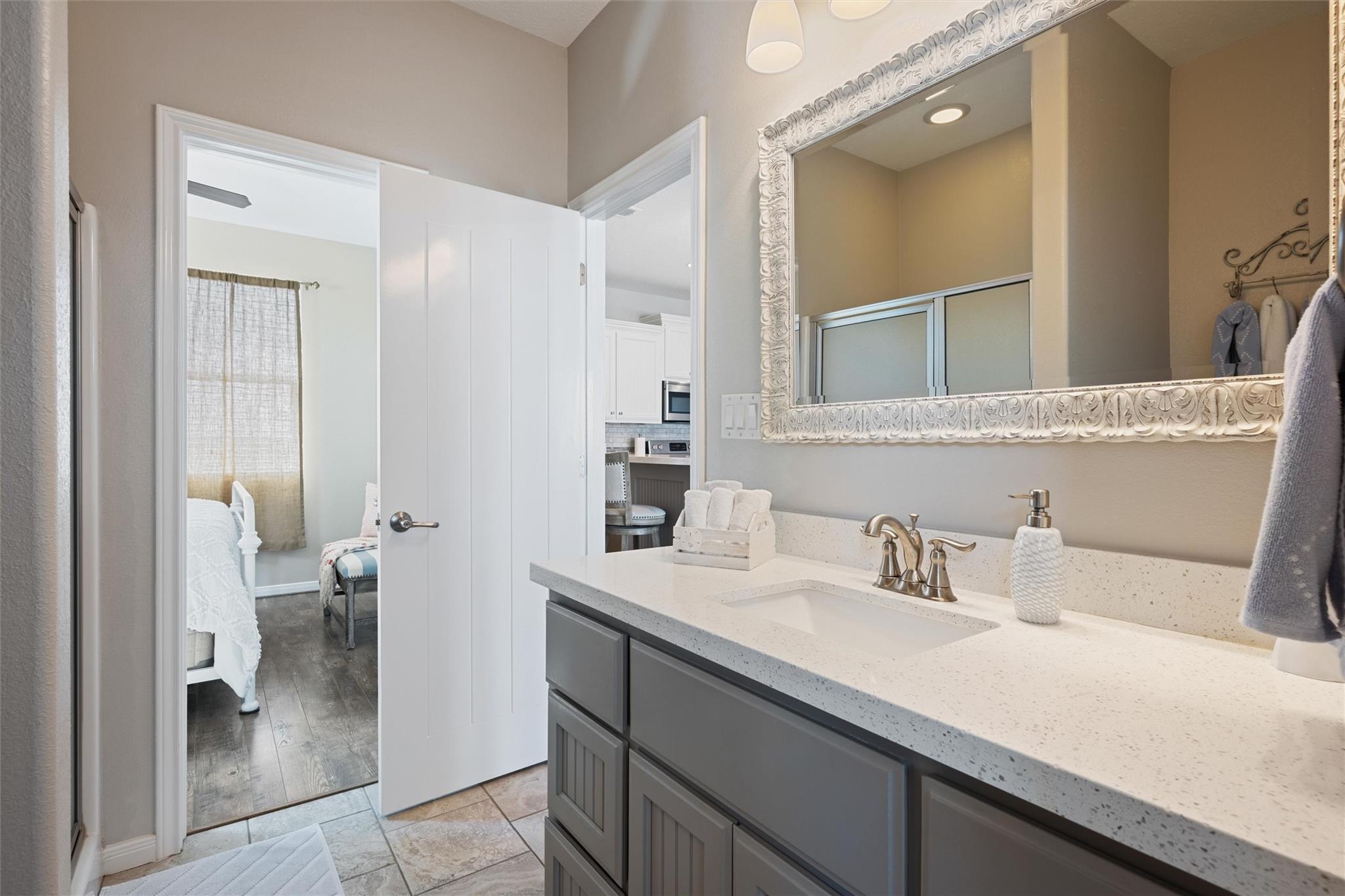 Bathroom vanity with gray cabinets, white countertop, ornate mirror, and doorway to a bedroom.