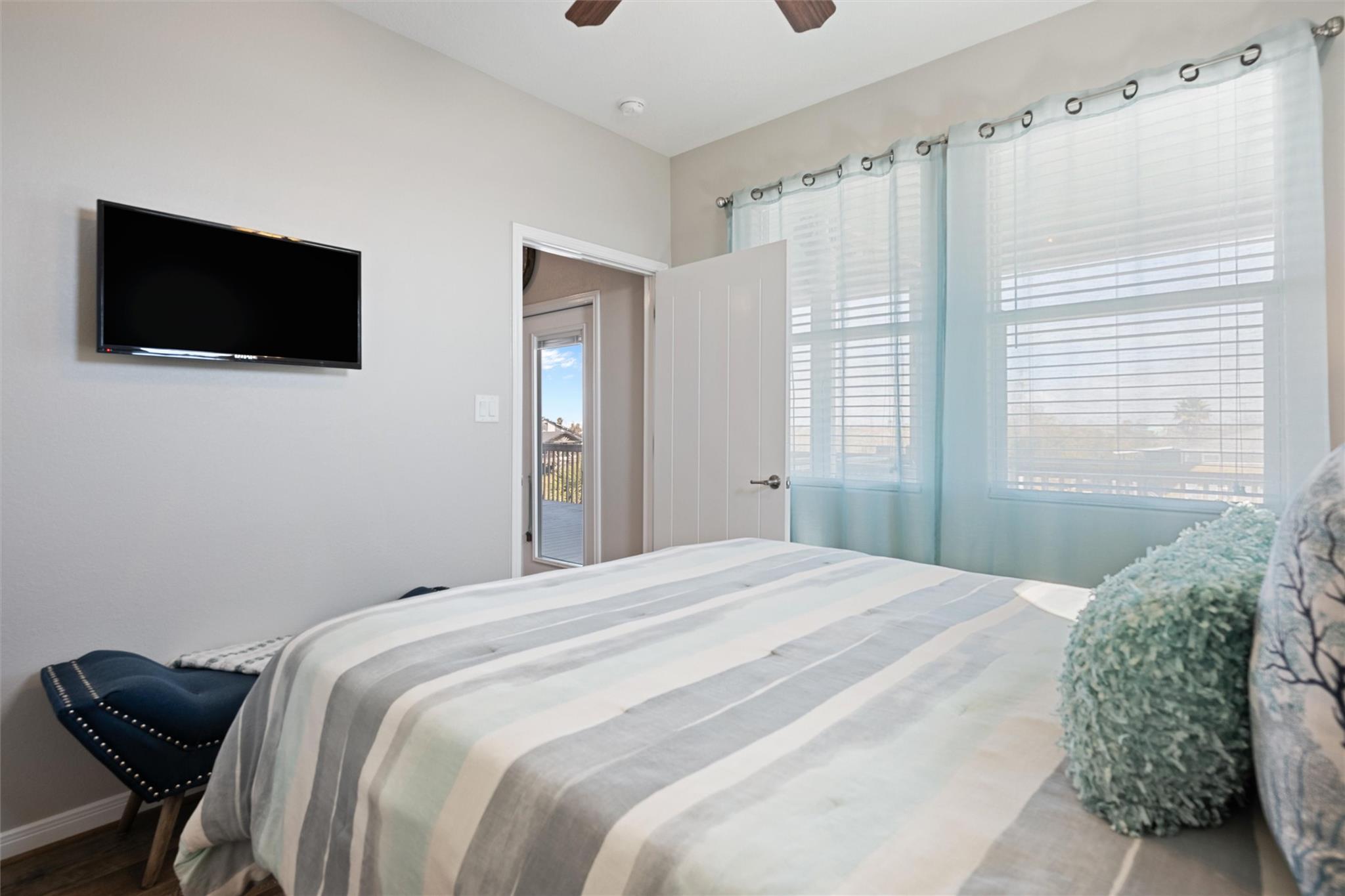 Bedroom with a striped bedspread, wall-mounted television, and pale blue curtains over wide windows.