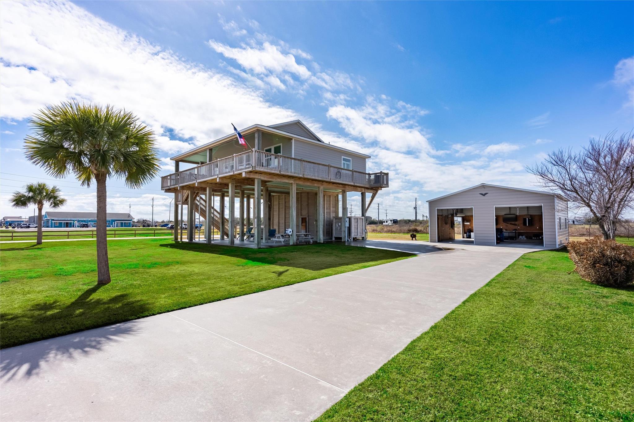 Raised house with wraparound deck and Texas flag beside a detached garage and wide driveway.