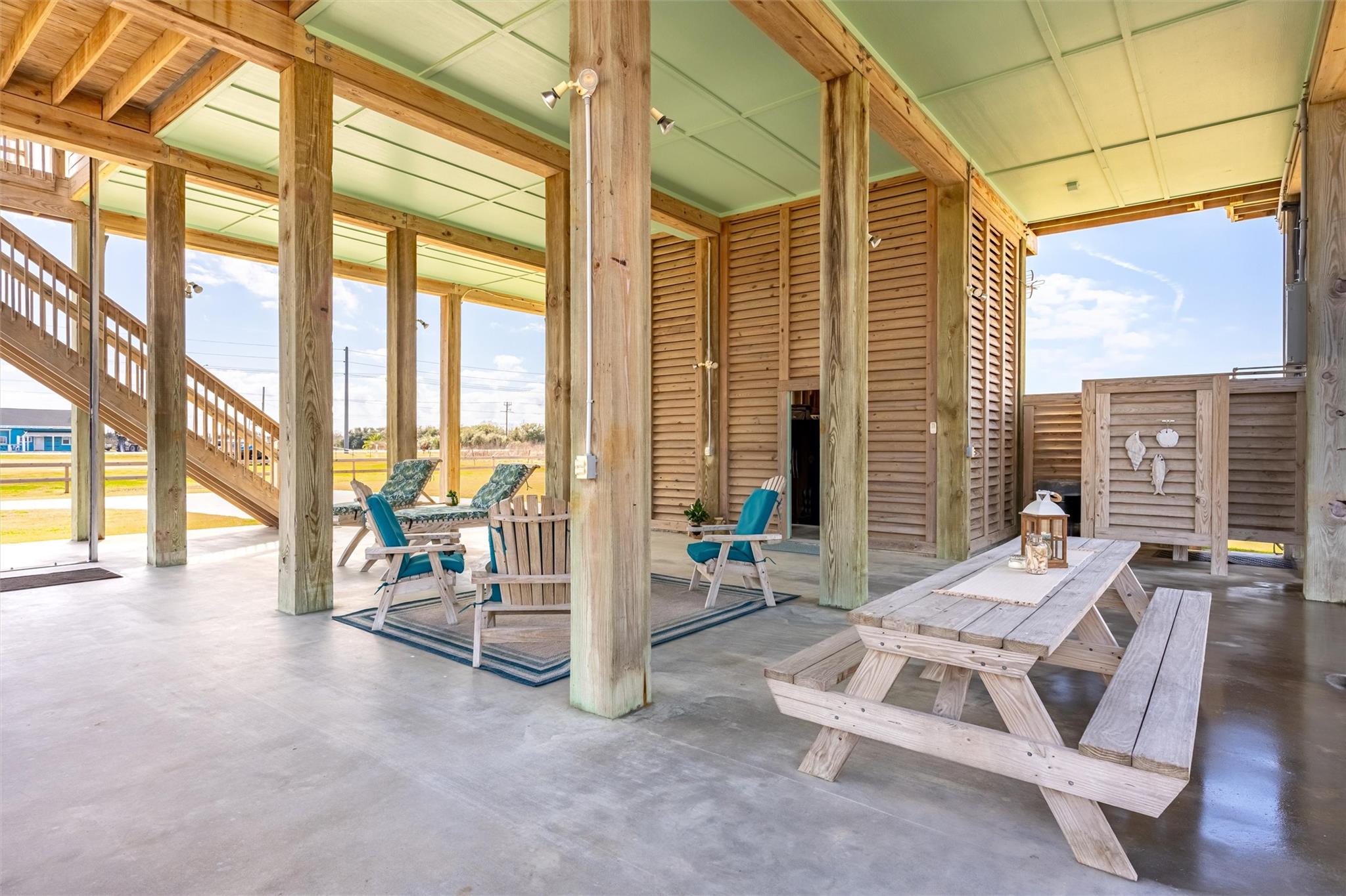Covered patio beneath a raised home with timber posts, lounge chairs, and a picnic table.