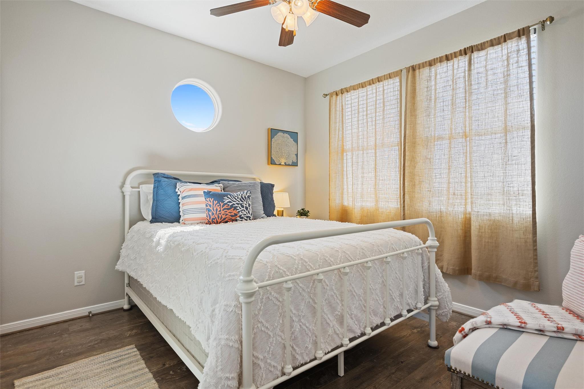 Staged bedroom with a white metal bed, blue accent pillows, and a round porthole-style window.