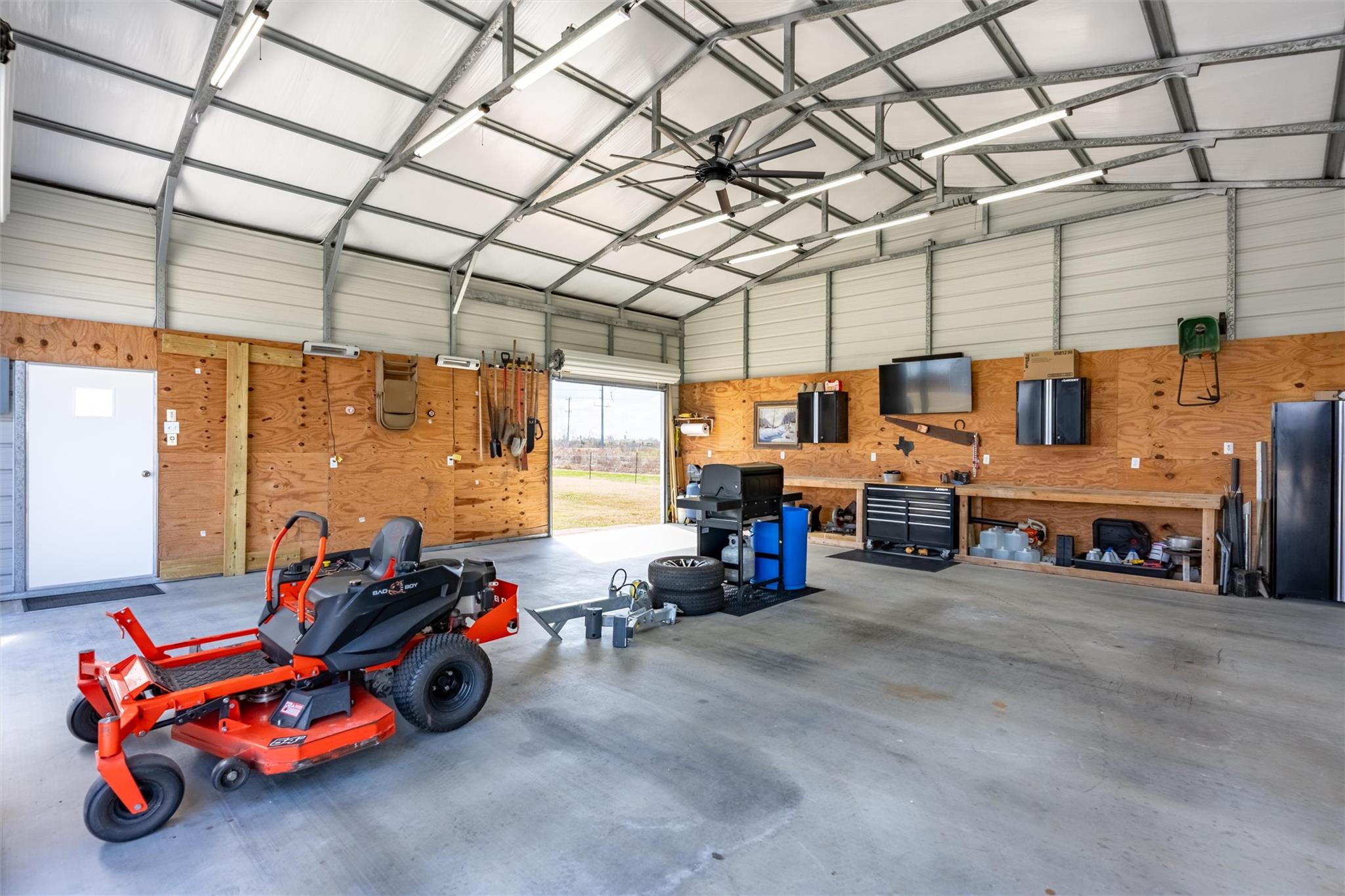 Spacious garage workshop with a red riding mower, wall-mounted tools, and a high metal-frame ceiling.