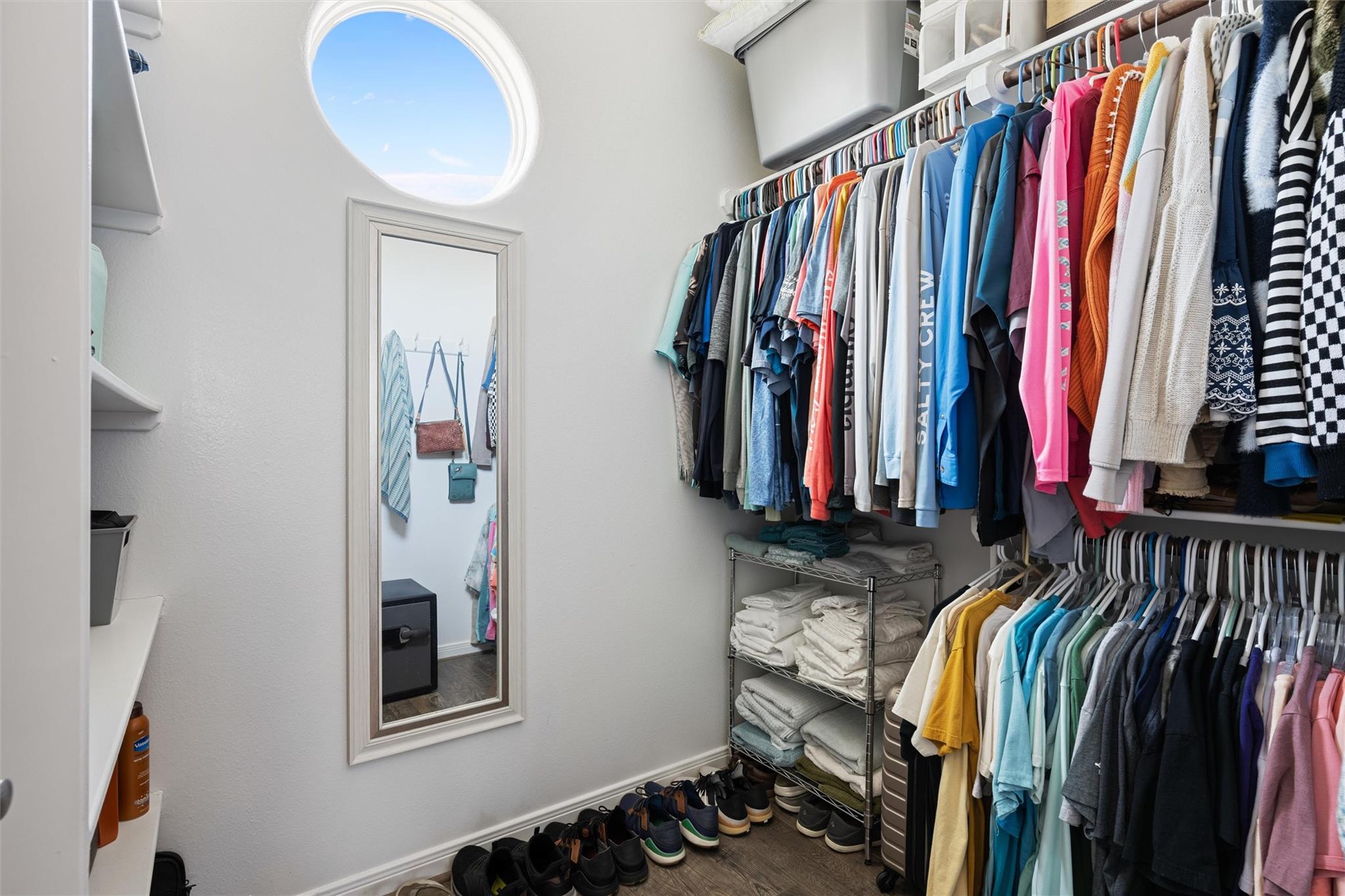Walk-in closet with colorful hanging clothes, neatly folded towels, rows of shoes, and a round window.