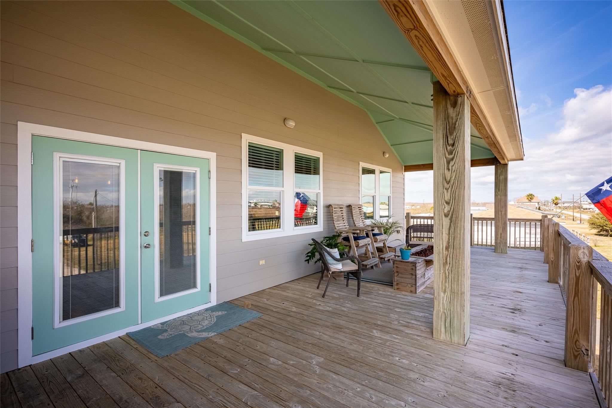 Covered wooden porch with aqua double doors, seating, timber posts, and a view over the coastal neighborhood.