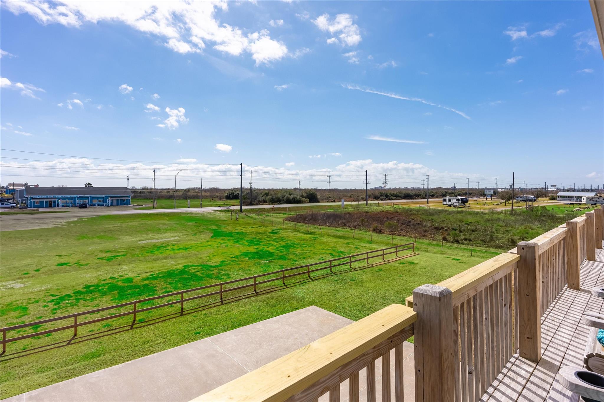 View from a wooden deck across a broad grassy lot toward a road and distant buildings.