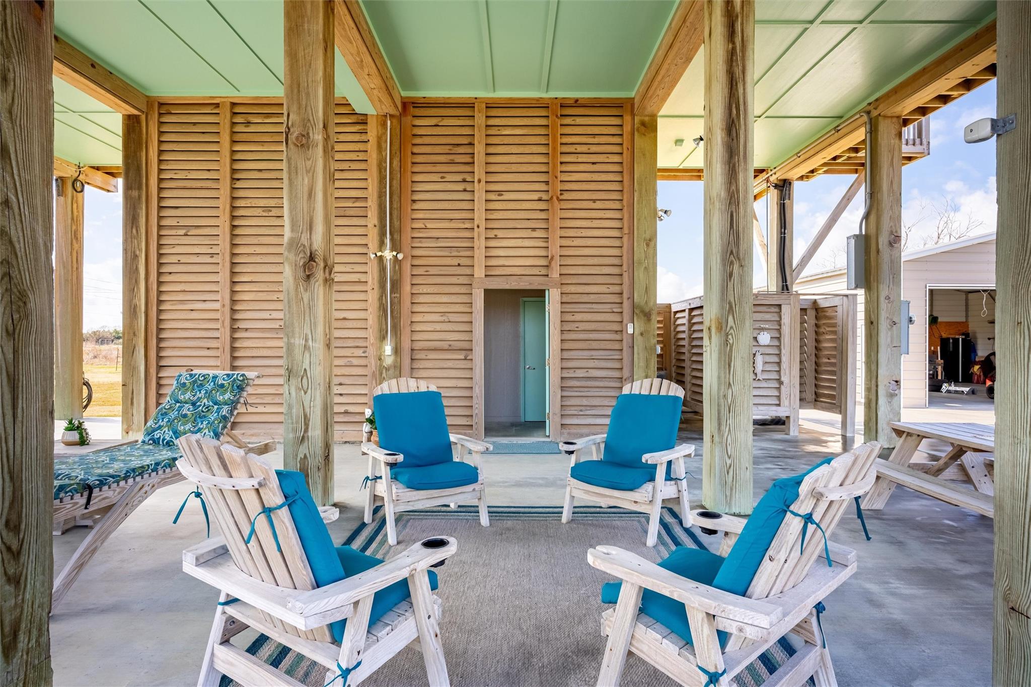 Covered timber patio furnished with blue-cushioned Adirondack chairs around a rug.