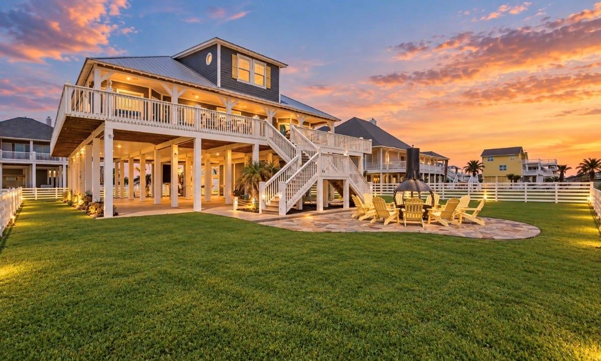 Elevated coastal home with wraparound decks, outdoor fire pit seating, and a broad lawn at sunset.