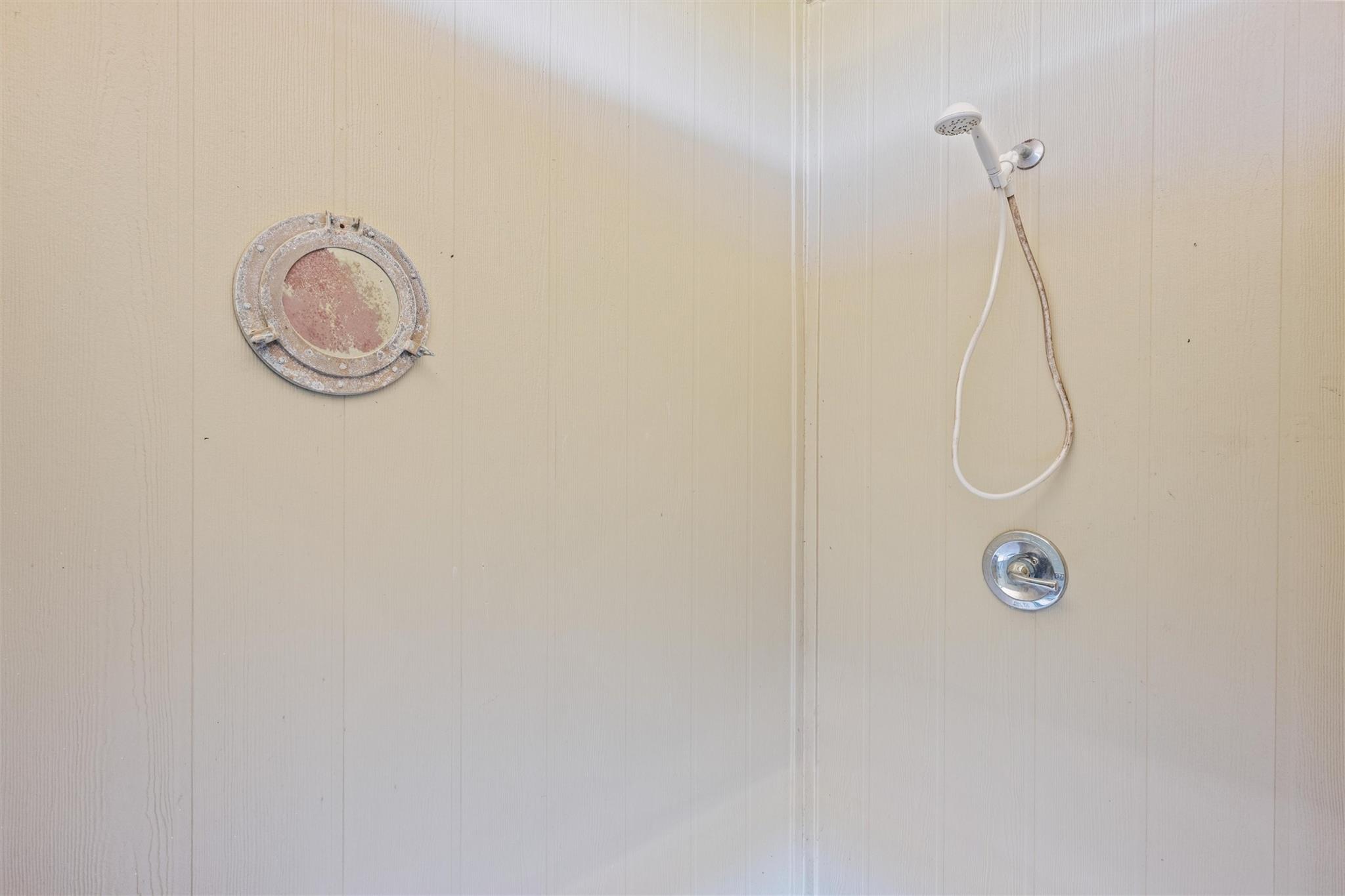 Outdoor shower on beige siding with a handheld showerhead and round porthole-style window.
