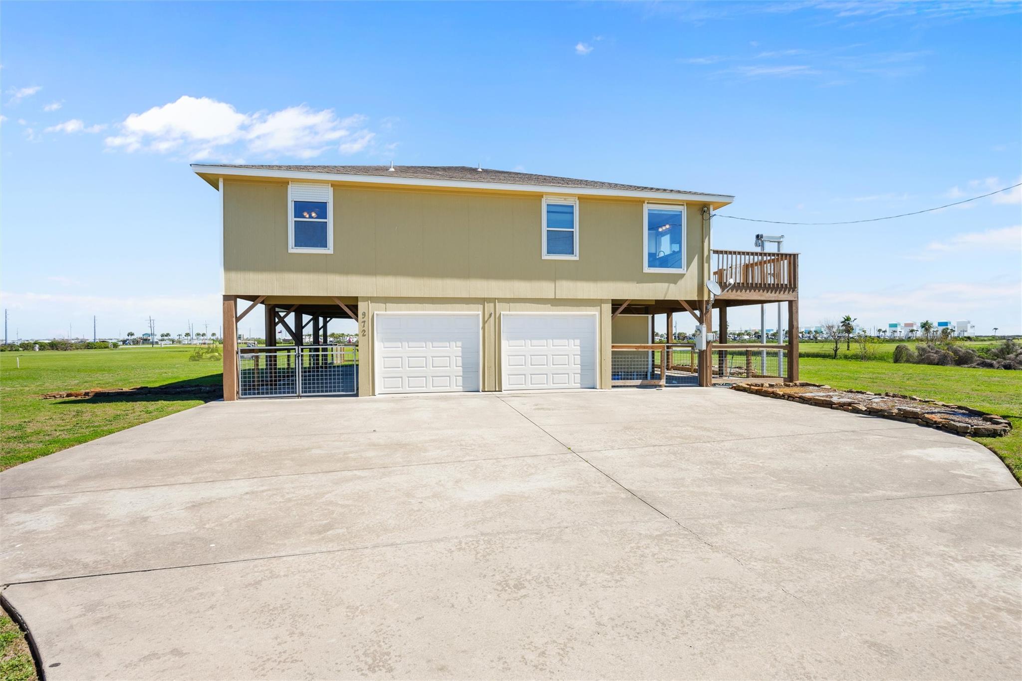 Raised tan house with two garage doors, a wooden side deck, and a broad concrete driveway.