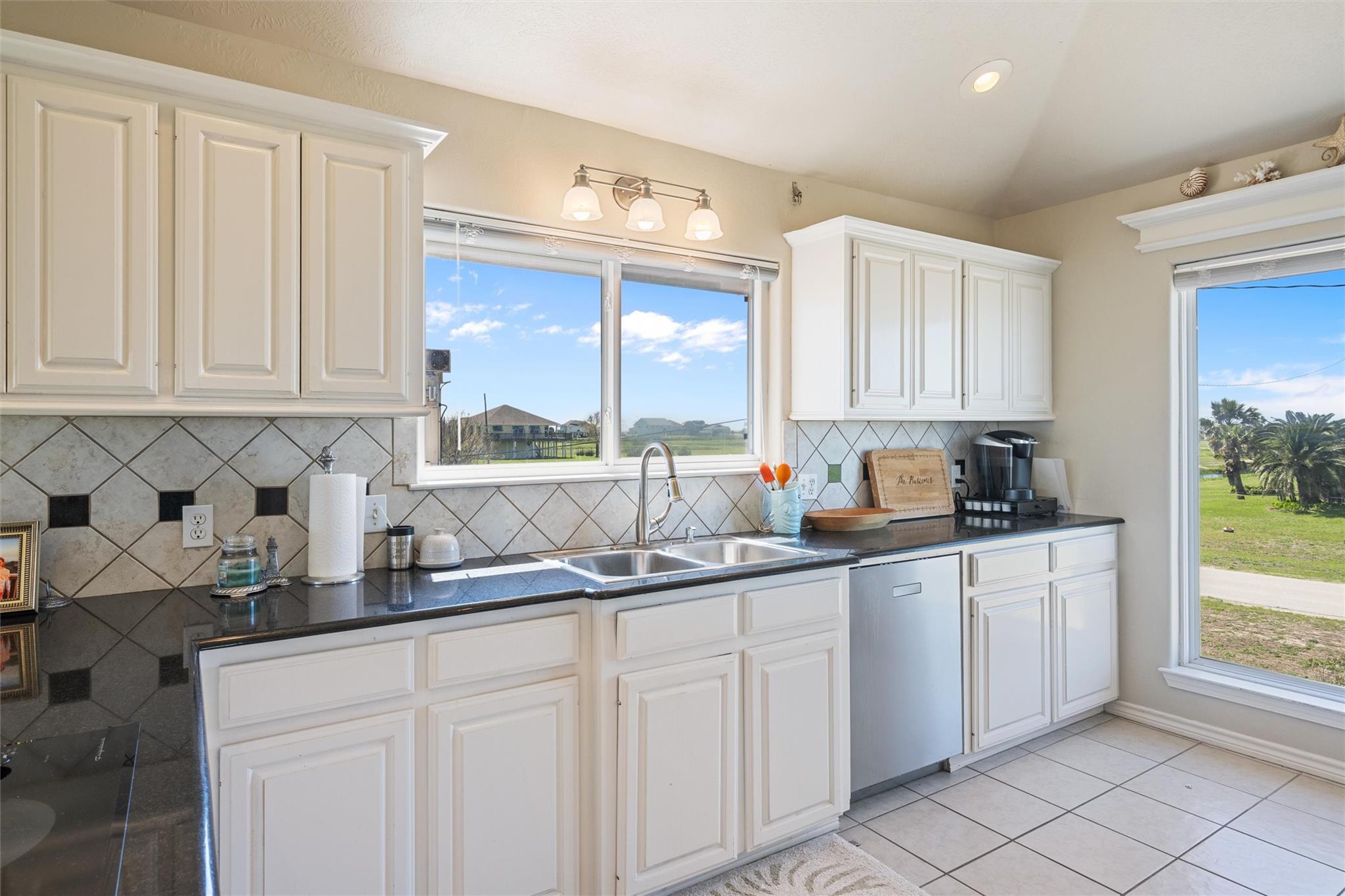 Kitchen with white cabinets, dark countertops, a diamond-pattern backsplash, and windows overlooking a grassy yard.