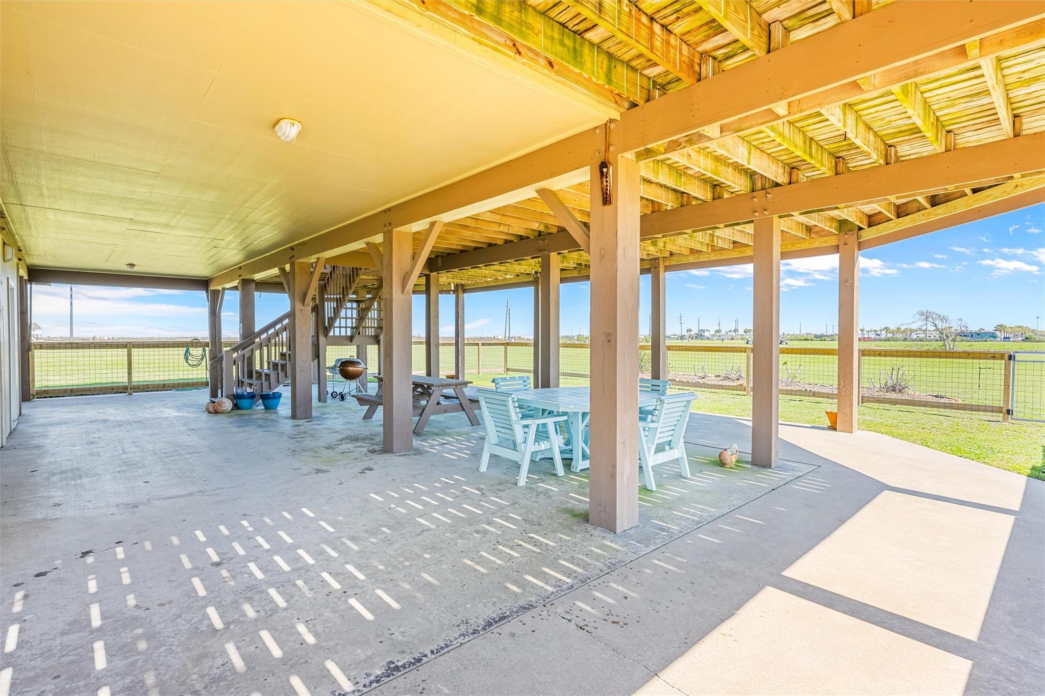 Covered patio with exposed beams, stairs, and an outdoor dining set overlooking a fenced lawn.