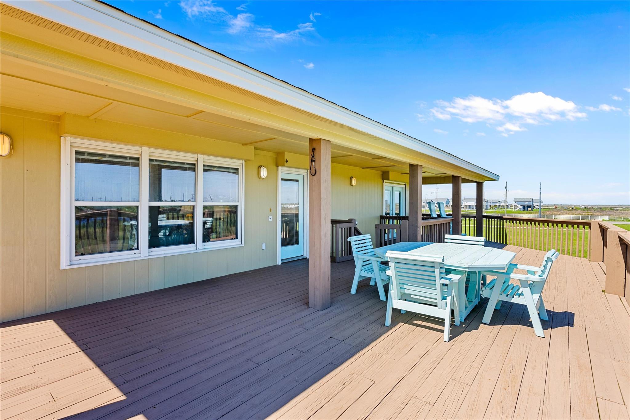 Covered wooden deck with white outdoor dining furniture beside a pale yellow home.