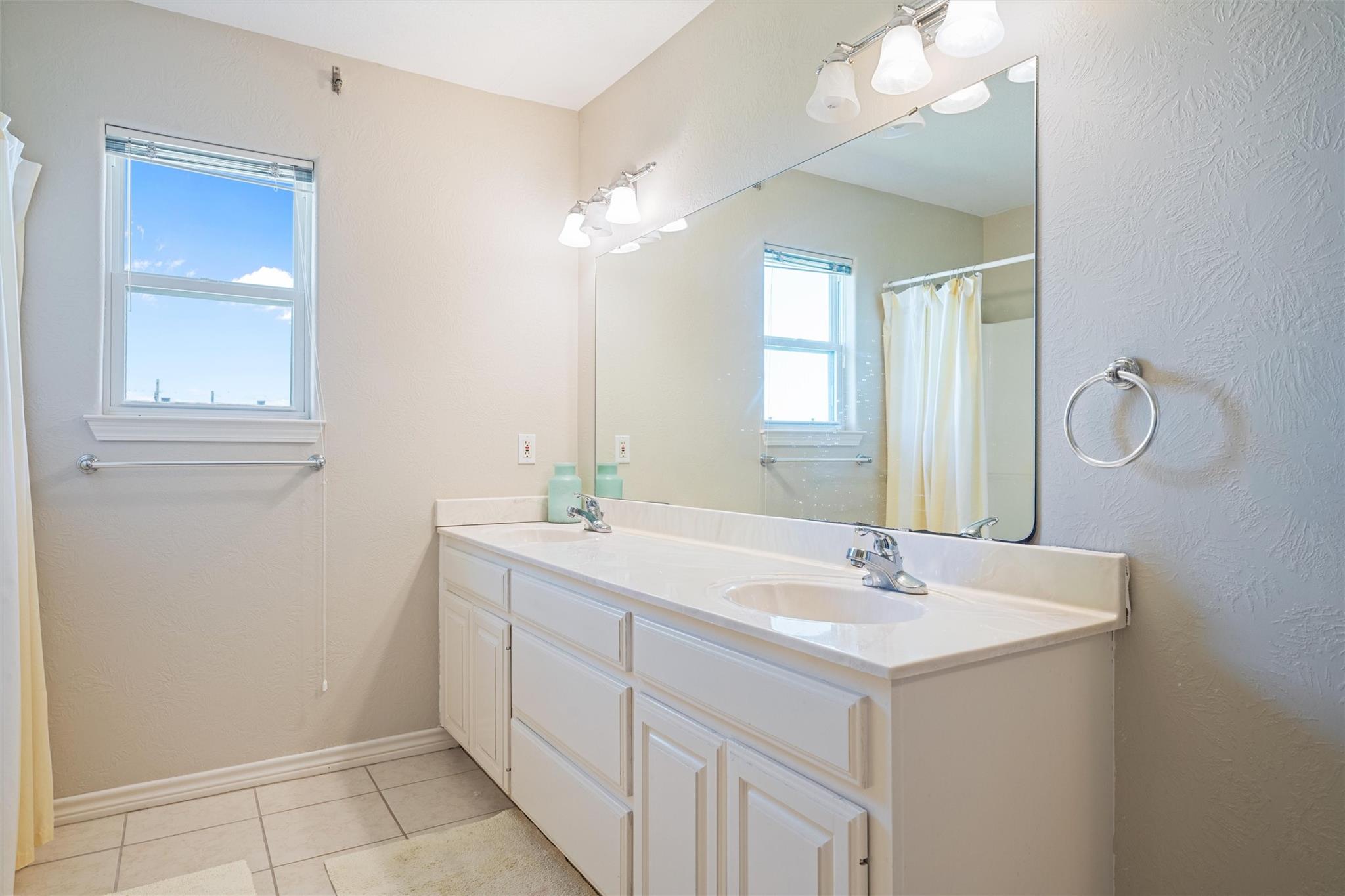 Bathroom with a long white double-sink vanity, oversized mirror, and window showing blue sky.