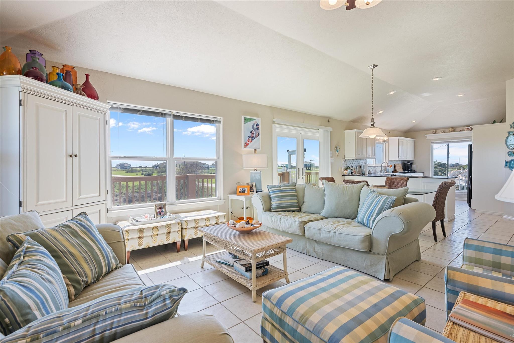 Furnished living room with vaulted ceiling, blue-striped seating, large windows, and an adjoining kitchen.