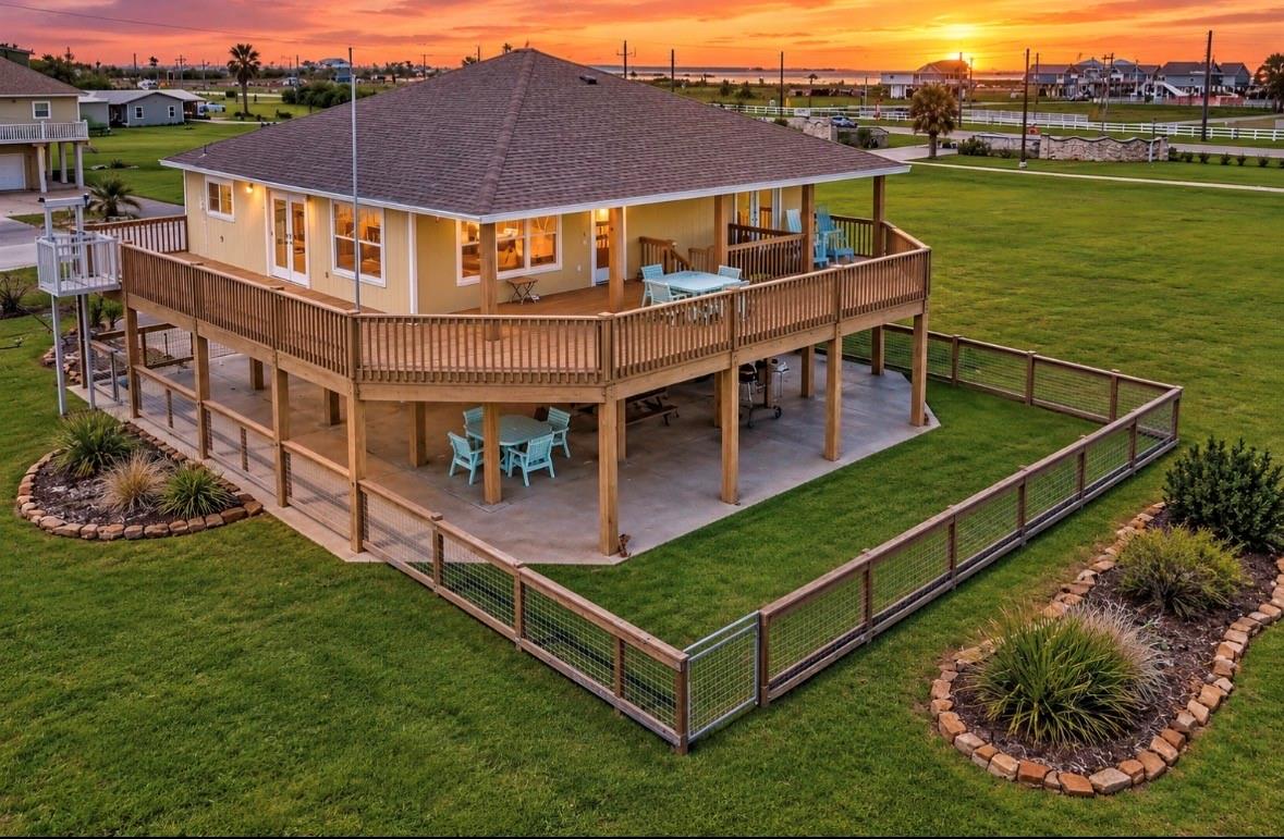 Elevated coastal home with a wraparound deck and fenced lawn at sunset.