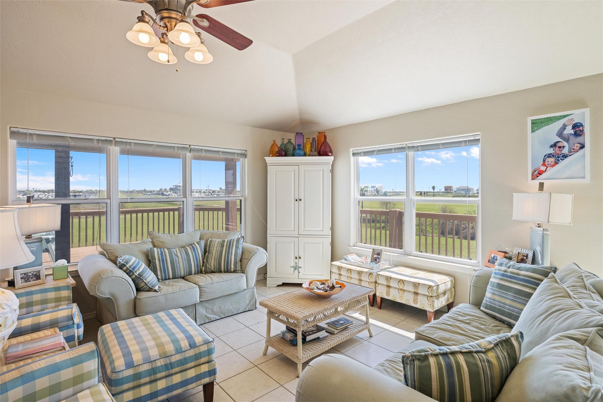 Furnished living room with large windows overlooking a deck and grassy coastal area.