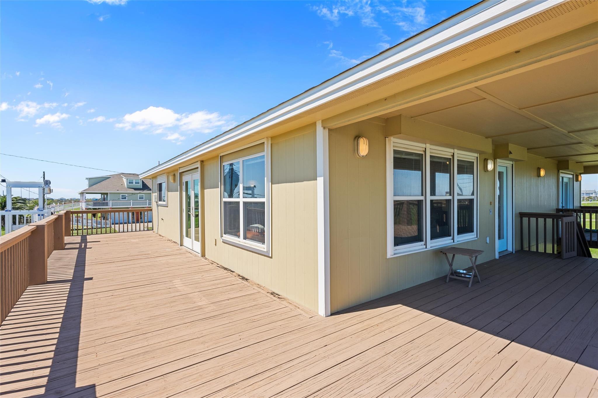 Tan home with a spacious wooden deck, covered patio, and water glimpsed beyond the railing.